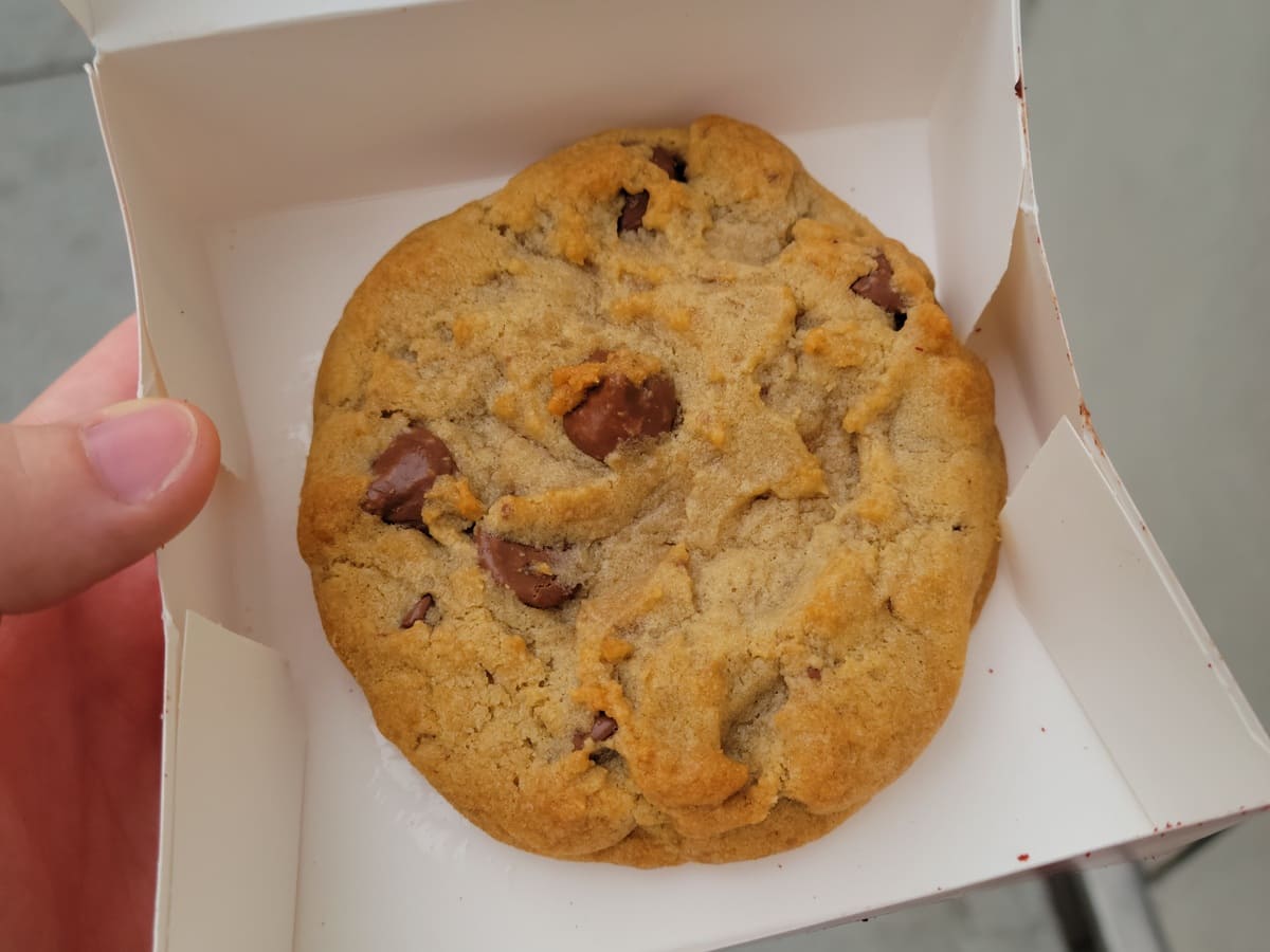 A person holding a chocolate chip cookie in a box, wondering how long do crumbl cookies last.